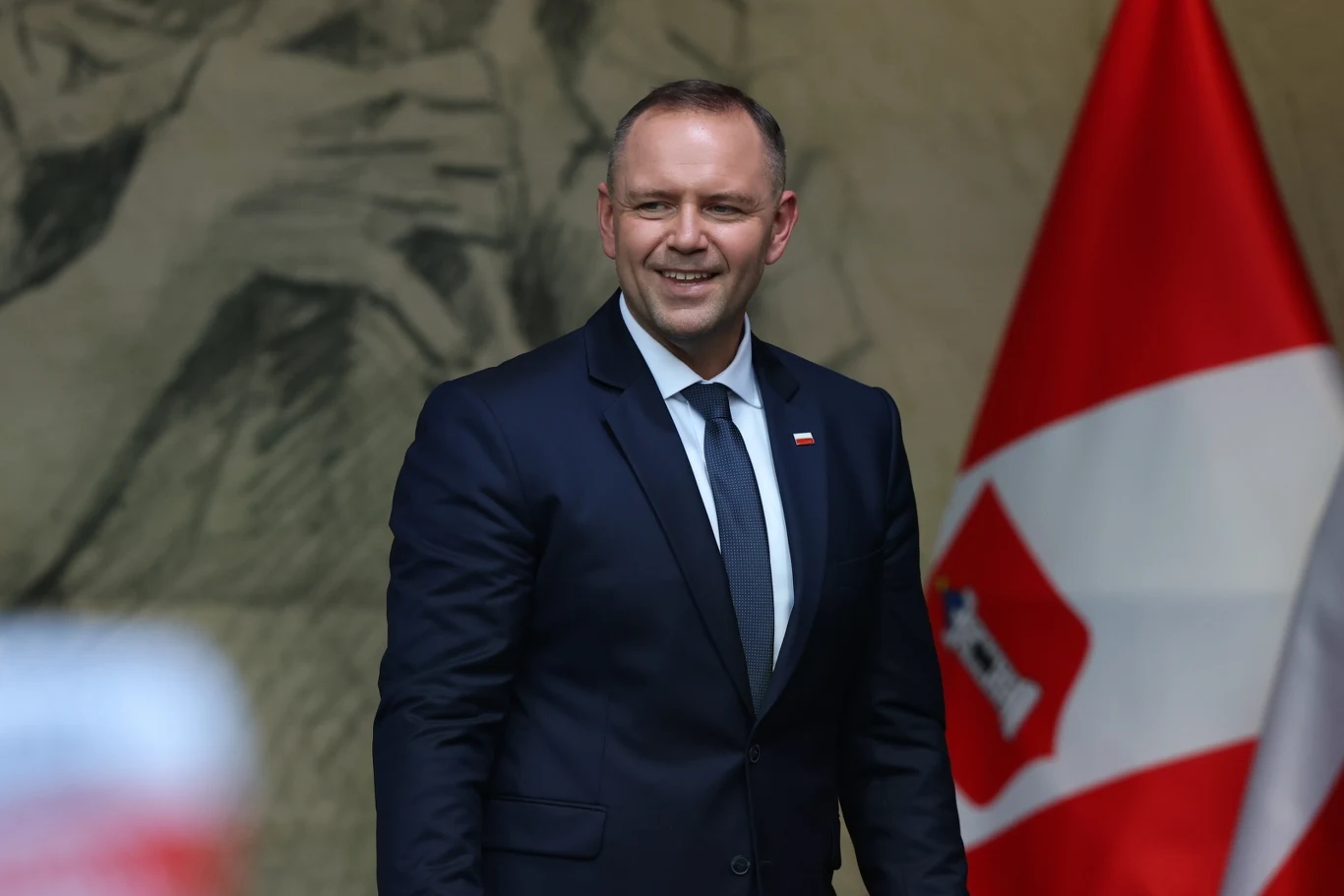 Prezydent Karol Nawrocki wziął udział w uroczystościach w Wieluniu Mężczyzna w granatowym garniturze z białą koszulą i krawatem stoi na tle flagi w biało-czerwonych kolorach oraz dużego szkicu na ścianie, uśmiechnięty patrzy w stronę aparatu.