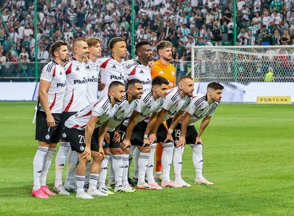 Legia Warszawa Grupa piłkarzy w biało-czarnych strojach z logo sponsora ustawia się do wspólnego zdjęcia na zielonej murawie stadionu, w tle widać liczną publiczność na trybunach oraz bramkę.