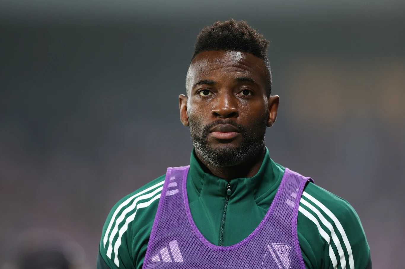 Mężczyzna w zielonej sportowej kurtce i fioletowej kamizelce stoi na stadionie, patrząc prosto w obiektyw; wyraz twarzy poważny, tło rozmyte.