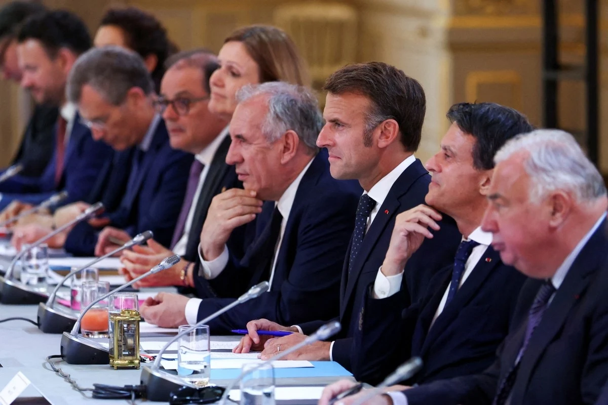 Grupa elegancko ubranych mężczyzn i kobiet siedzi przy długim stole konferencyjnym, zajęci rozmową lub wymianą uwag. W tle widać złocone dekoracje, co sugeruje prestiżowe wnętrze o pałacowym charakterze.