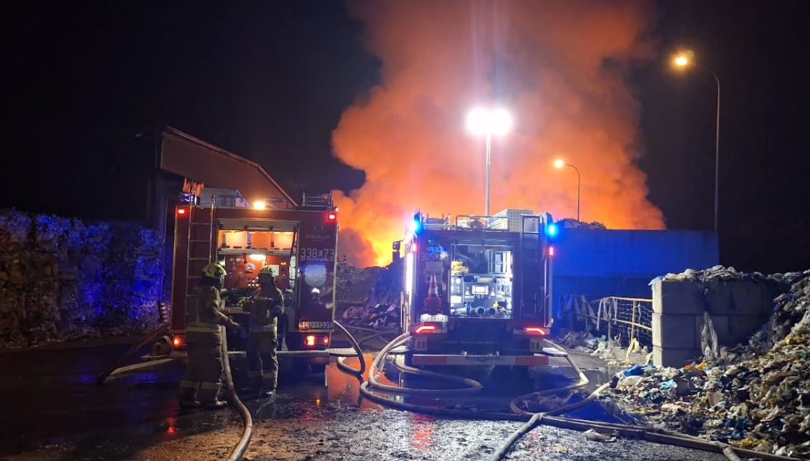 Trwa akcja gaśnicza w Bełżycach. Z ogniem walczy ponad 100 strażaków Dwie strażackie wozy ratownicze stojące przed dużym pożarem, z intensywnie unoszącym się pomarańczowym dymem na tle nocnego nieba, otoczone stertami śmieci i odpadów, strażacy prowadzą akcję gaśniczą.