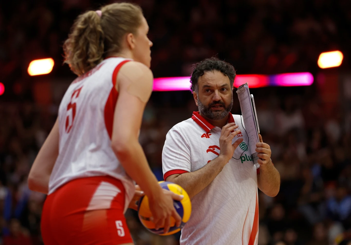Trener drużyny siatkówki w białoczerwonym stroju rozmawia z zawodniczką trzymającą piłkę, w tle rozmyte sylwetki kibiców oraz rozświetlona hala sportowa.