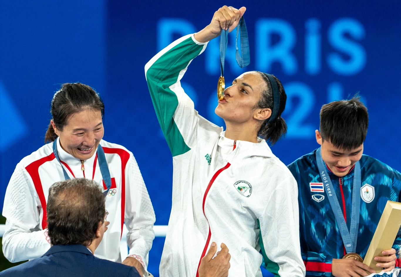 Sportowcy stojący na podium, kobieta w białym stroju całuje złoty medal, obok znajdują się dwie osoby z medalami i dyplomami, w tle napis 'Paris 2024'.