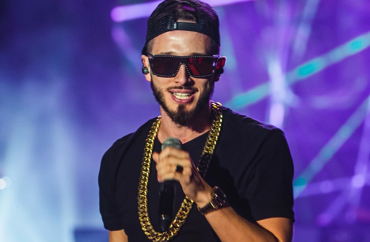 Bogumił "Boogie" Romanowski Mężczyzna w czarnej koszulce, z brodą, noszący okulary przeciwsłoneczne, złoty łańcuch i czapkę z daszkiem, śpiewa do mikrofonu na scenie z kolorowym oświetleniem w tle.