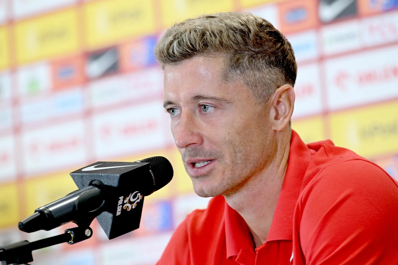 Mężczyzna w czerwonej koszulce udzielający wywiadu podczas konferencji prasowej, na tle ściany z logotypami sponsorów i partnerów sportowych.