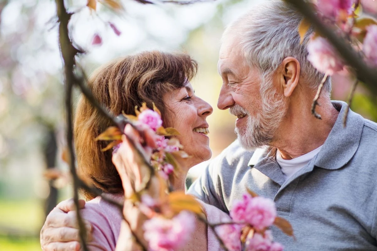 Para starszych ludzi przytulająca się wśród kwitnących gałęzi, patrzących sobie w oczy i uśmiechających się z czułością