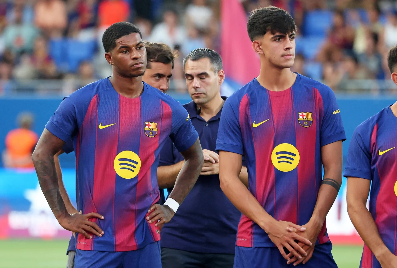 Dwóch młodych piłkarzy w strojach FC Barcelony stoi z poważnymi minami podczas oficjalnego wydarzenia, w tle inne osoby i widownia stadionowa.
