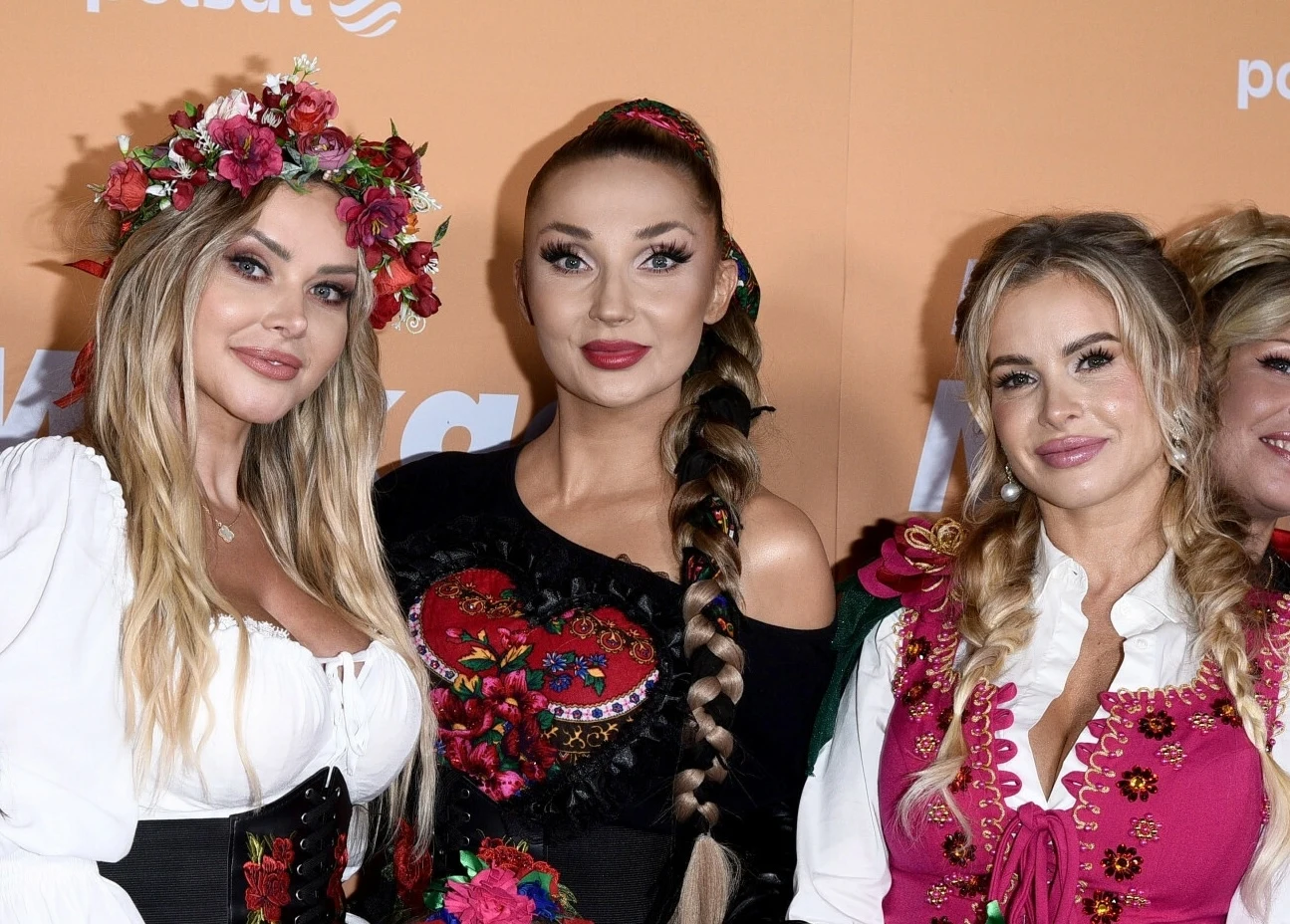 Ola Ciupa, Cleo i Paulina Tomala spotkały się ponownie Trzy kobiety ubrane w tradycyjne stroje ludowe o bogatych zdobieniach, z kwiatowymi motywami i kwiatowymi wiankami na głowach, stojące razem na tle pomarańczowej ścianki.