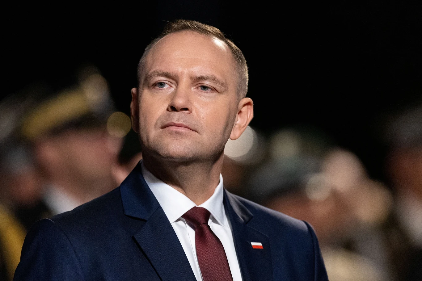 Prezydent Karol Nawrocki złoży wizytę w USA. Nie będzie przedstawiciela polskiego MSZ Mężczyzna w ciemnym garniturze i bordowym krawacie patrzący poważnie w bok, na tle rozmytej grupy osób w mundurach, z polską flagą przypiętą do klapy marynarki.