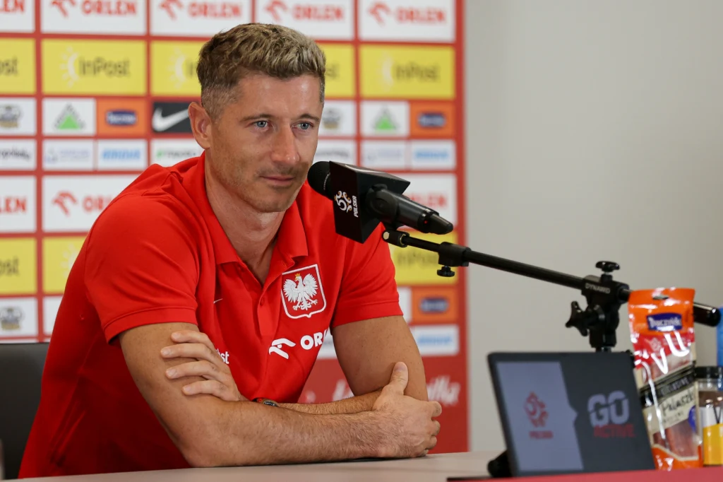 Robert Lewandowski na konferencji Mężczyzna w czerwonej koszulce z godłem Polski udziela wywiadu podczas konferencji prasowej, siedząc przy stole z mikrofonem, przed ścianą z logo sponsorów.