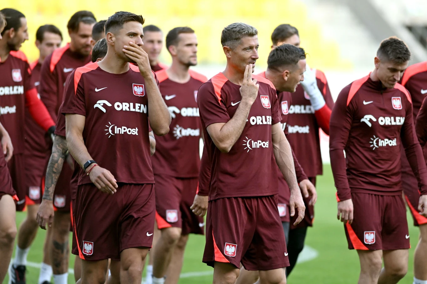 Polscy piłkarze drużyny narodowej w bordowych strojach sportowych podczas treningu na boisku, część z nich przechadza się po murawie, a w tle widać resztę zespołu.