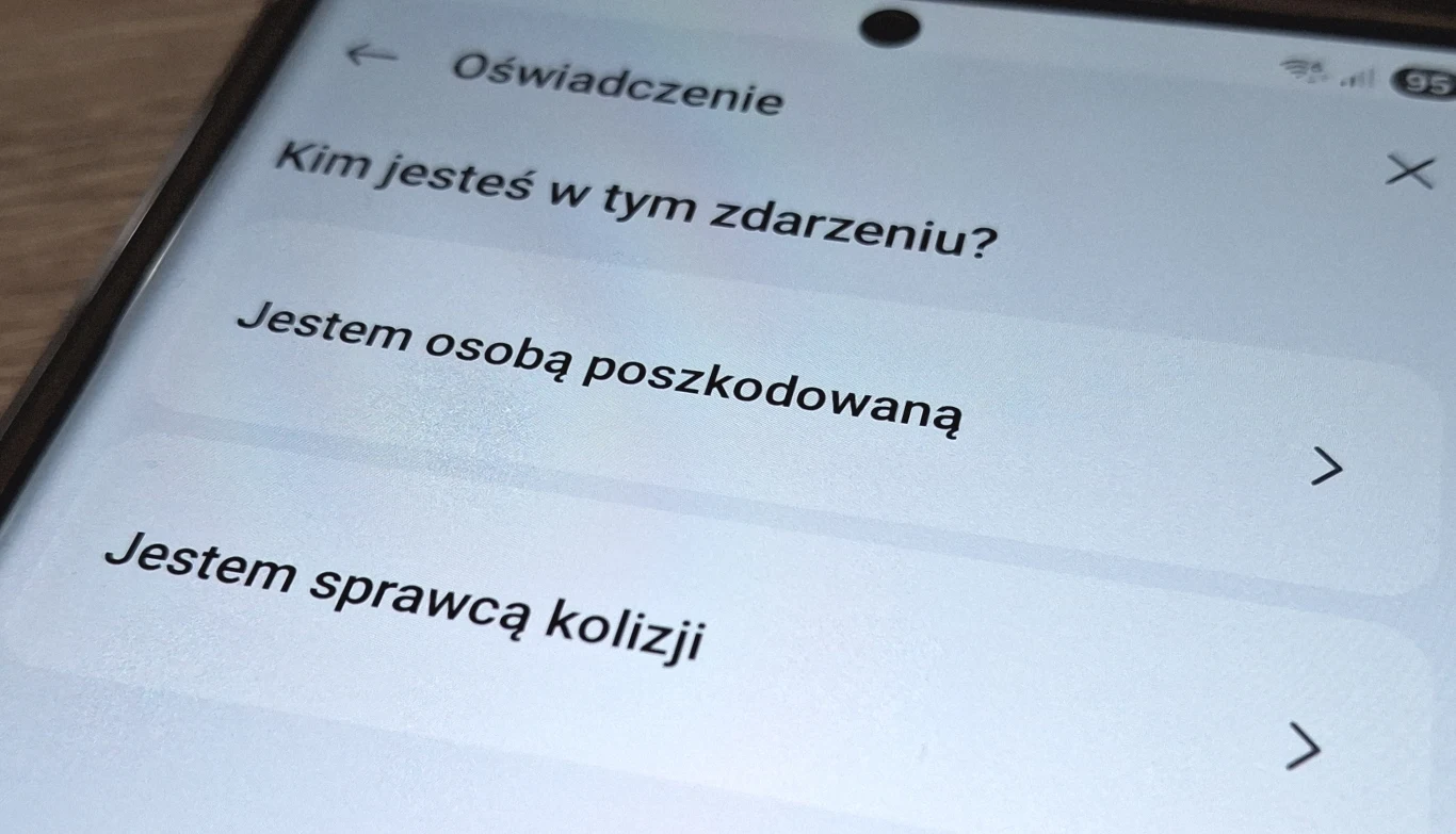 Ekran telefonu z aplikacją, na której widoczne są opcje wyboru: 'Jestem osobą poszkodowaną' i 'Jestem sprawcą kolizji', dotyczące zgłoszenia zdarzenia.