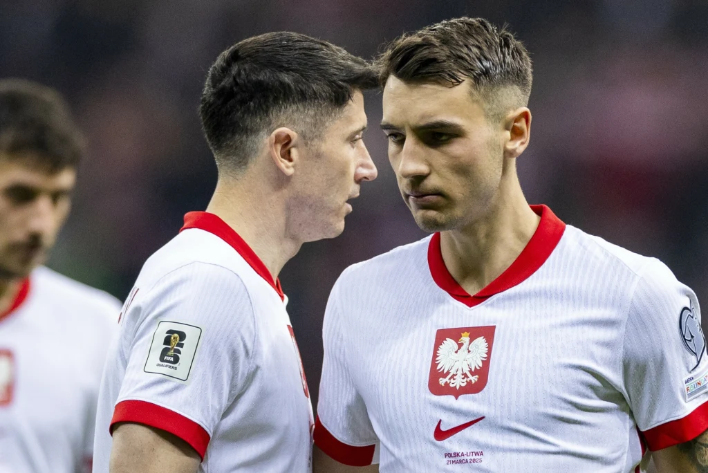 Jakub Kiwior i Robert Lewandowski w reprezentacji Polski Dwóch piłkarzy w białych koszulkach z godłem Polski stoi obok siebie, jeden z nich wydaje się mówić do drugiego, na twarzach skupienie i powaga, w tle widoczny jest rozmyty stadion.