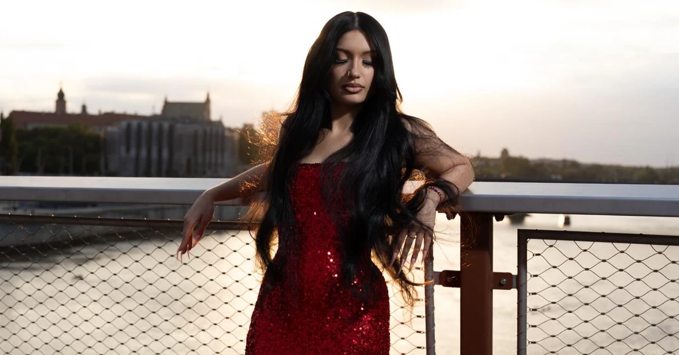 Viki Gabor w rozmowie z Interią Muzyką Kobieta o długich czarnych włosach w czerwonej cekinowej sukni opiera się o poręcz mostu z widokiem na rzekę i zabudowania w tle, światło zachodzącego słońca delikatnie podkreśla jej sylwetkę.