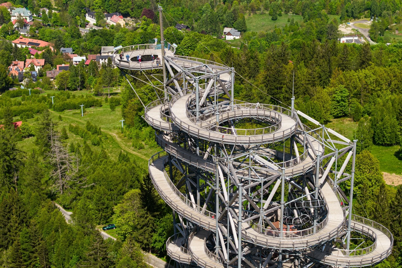 Stalowa konstrukcja ścieżki w koronach drzew wijąca się spiralnie ku górze, otoczona zielonym lasem, z widokiem na pobliskie zabudowania i rozległe tereny zielone.