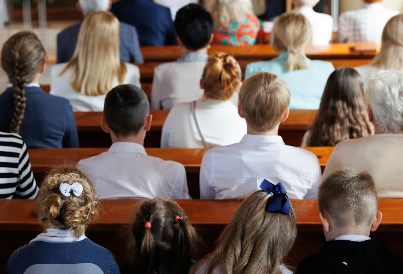 Światowy Dzień Misyjny 2025. Co to za święto? Dzieci i dorośli siedzą w ławkach, odwróceni tyłem, skupieni na wydarzeniu odbywającym się przed nimi, nieokreślonym z perspektywy widza. Uczesane dziewczynki z kokardami, chłopcy w białych koszulach, atmosfera powagi i skupienia.