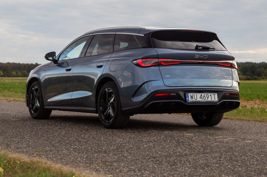 Elektryczny samochód BYD Dolphin w metalicznym kolorze niebieskim stoi na asfaltowej drodze, widok z tyłu, czarne felgi, widoczne tablice rejestracyjne i zielone pola w tle.