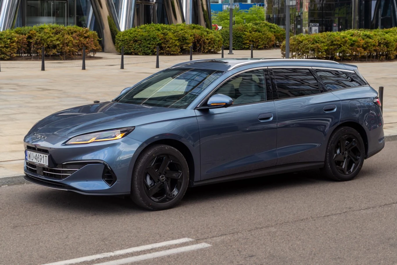 Nowoczesny szary samochód osobowy typu kombi zaparkowany przy ulicy w mieście, otoczony niskimi żywopłotami oraz nowoczesną architekturą ze szkła i stali w tle.