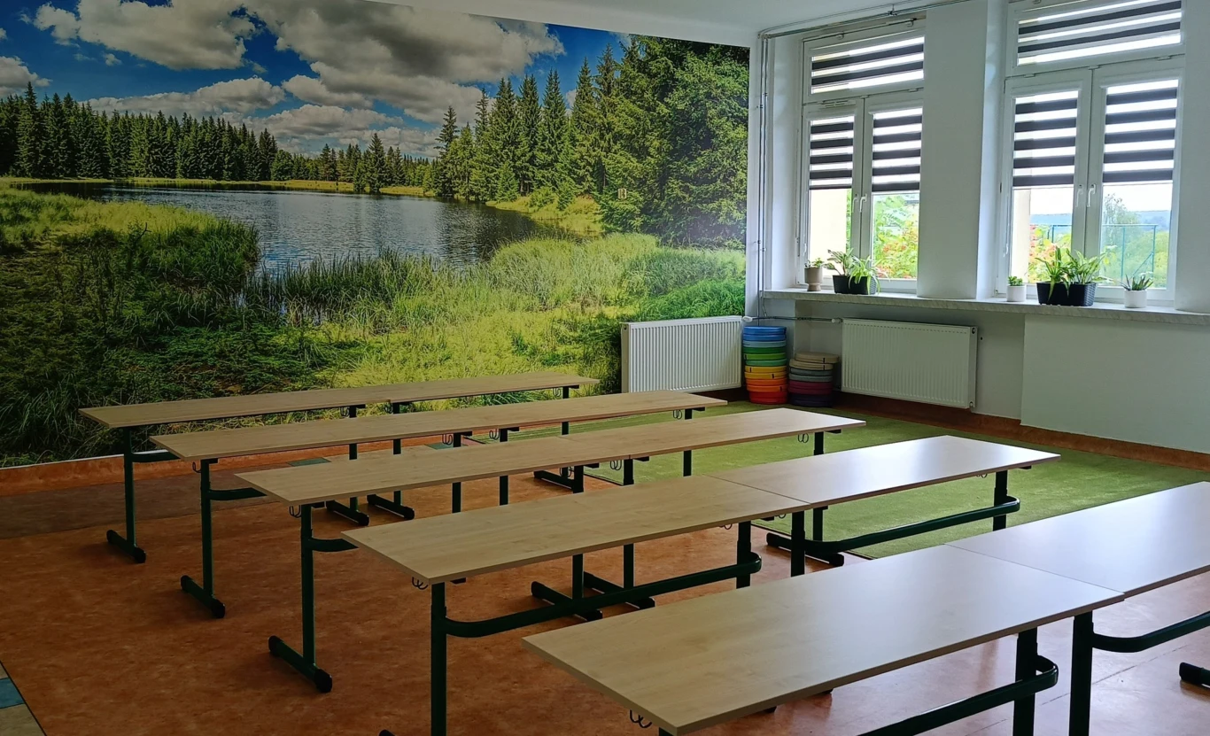Szkoła w Starachowicach. Nagranie z rozpoczęcia roku szkolnego wywołało kontrowersje Pusta sala lekcyjna z kilkoma rzędami drewnianych ławek, na ścianie fototapeta przedstawiająca jezioro i las, pod dużymi oknami stoją doniczki z roślinami i kolorowe siedzenia w rogu.
