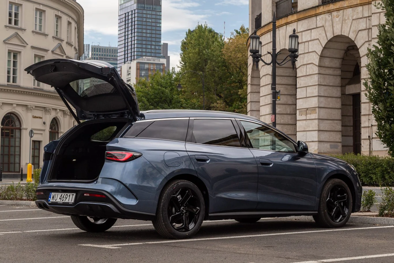 Nowoczesny, niebieski samochód kombi zaparkowany na tle miejskich budynków z otwartym bagażnikiem, widoczne czarne felgi oraz przeszklone fragmenty karoserii.