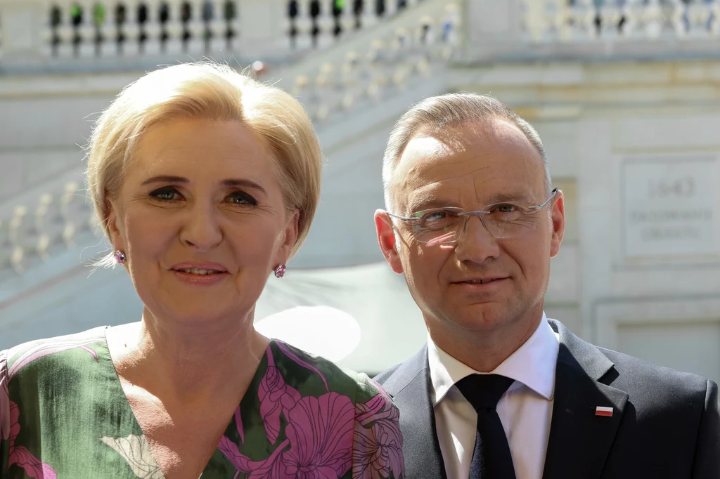 Przeprowadzka z Warszawy do Krakowa była dla Agaty i Andrzeja Dudów wyzwaniem Dwie elegancko ubrane osoby stoją blisko siebie na tle jasnych schodów i balustrady budynku w słonecznym oświetleniu, kobieta w sukience z kwiatowym motywem, mężczyzna w garniturze z przypiętą flagą Polski.