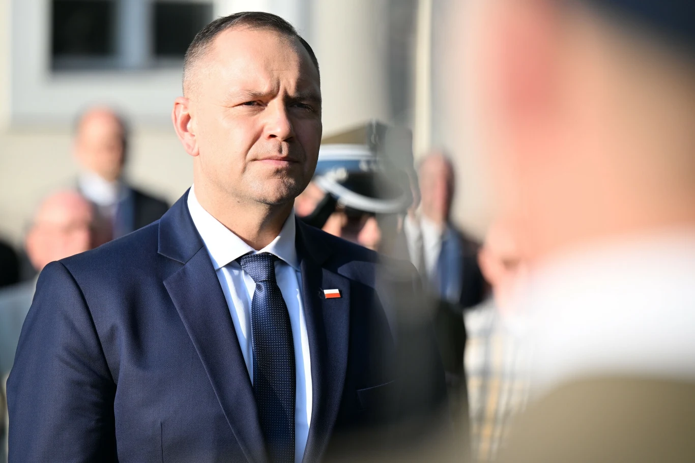 Mężczyzna w garniturze z przypiętą biało-czerwoną flagą stoi na zewnątrz, w tle rozmyte postaci i budynek.