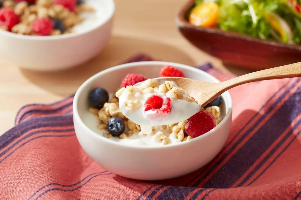 Jogurt naturalny to świetny pomysł na śniadanie. W towarzystwie owoców jagodowych skutecznie poskromi wysokie ciśnienie Miska jogurtu z musli, malinami i borówkami, na łyżce wyeksponowany owoc, w tle fragment sałatki i druga miseczka, całość ustawiona na tkaninie w prążki.