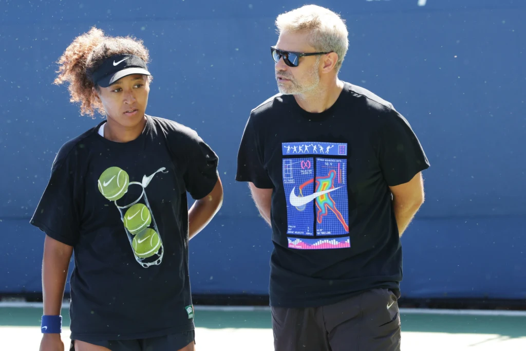 Naomi Osaka i Tomasz Wiktorowski Dwoje ludzi ubranych w sportowe koszulki stoi obok siebie na tle niebieskiego boiska tenisowego, kobieta po lewej stronie ma na koszulce nadrukowane piłki tenisowe, mężczyzna po prawej stronie nosi okulary przeciwsłoneczne i czarną koszulkę z kolorowym...