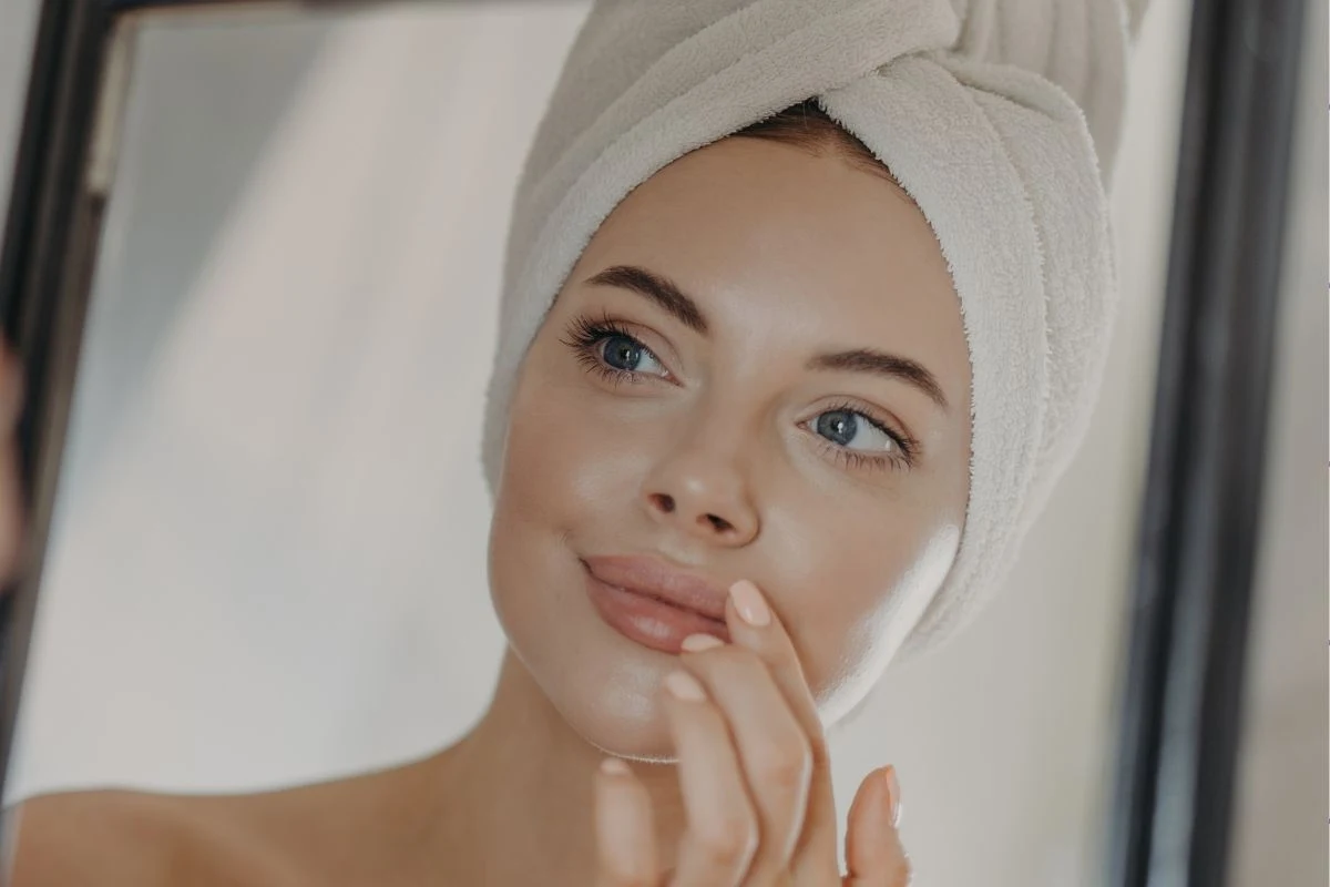 Zmęczona i opuchnięta cera? Ten trik działa natychmiast Młoda kobieta z ręcznikiem owiniętym na głowie stoi przed lustrem i delikatnie dotyka twarzy, prezentując zadbaną cerę i naturalny makijaż.