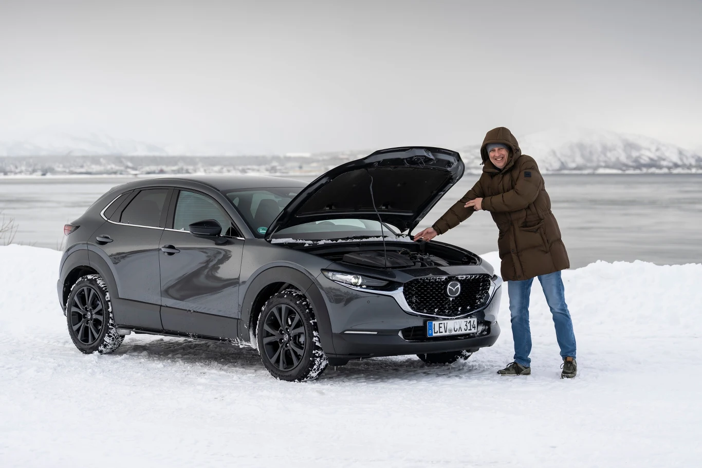 Osoba w kurtce zimowej stoi przy czarnym samochodzie marki Mazda zaparkowanym na śniegu, z otwartą maską silnika, w tle widoczne zimowe krajobrazy i zamarznięta woda.
