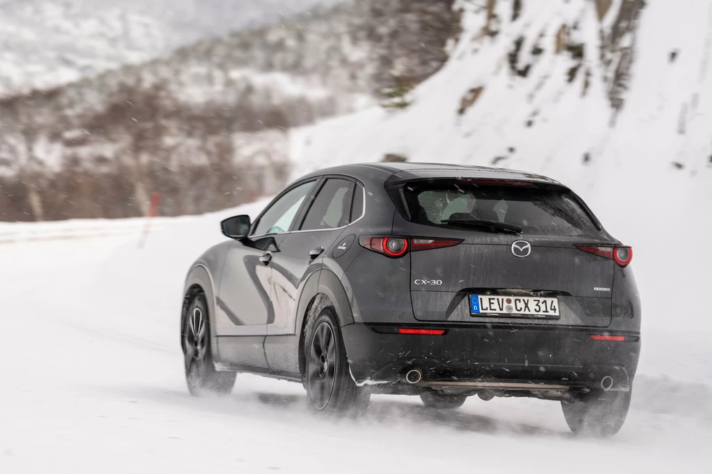 Zawieszenie Mazdy CX-30 na pokrytej lodem drodze pracowało bardzo cicho. Szary samochód marki Mazda CX-30 jedzie zaśnieżoną drogą pośród górskich krajobrazów, ślady kół widoczne w świeżym śniegu, w tle ośnieżone wzgórza i skały.