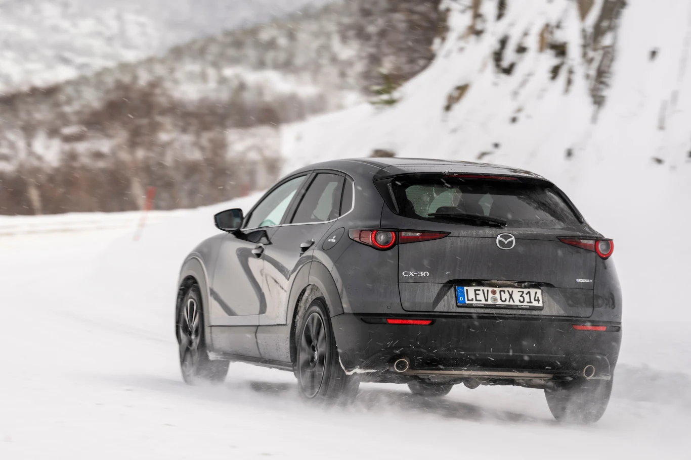 Szary samochód marki Mazda CX-30 jedzie zaśnieżoną drogą pośród górskich krajobrazów, ślady kół widoczne w świeżym śniegu, w tle ośnieżone wzgórza i skały.