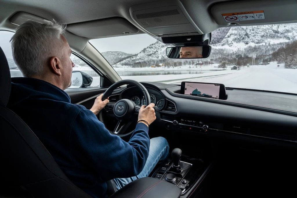 Mazda CX-30 prowadzi się nieźle, ale w trudnych warunkach chciałoby się czegoś więcej Mężczyzna o siwych włosach prowadzi samochód, jadąc zimową drogą przez góry, wewnątrz pojazdu widać deskę rozdzielczą, kierownicę oraz zamontowaną nawigację GPS z mapą.
