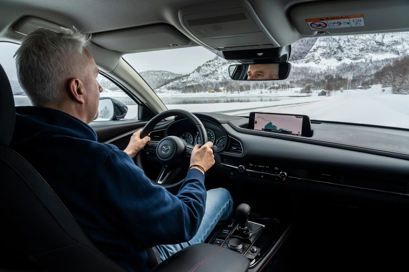 Mężczyzna o siwych włosach prowadzi samochód, jadąc zimową drogą przez góry, wewnątrz pojazdu widać deskę rozdzielczą, kierownicę oraz zamontowaną nawigację GPS z mapą.