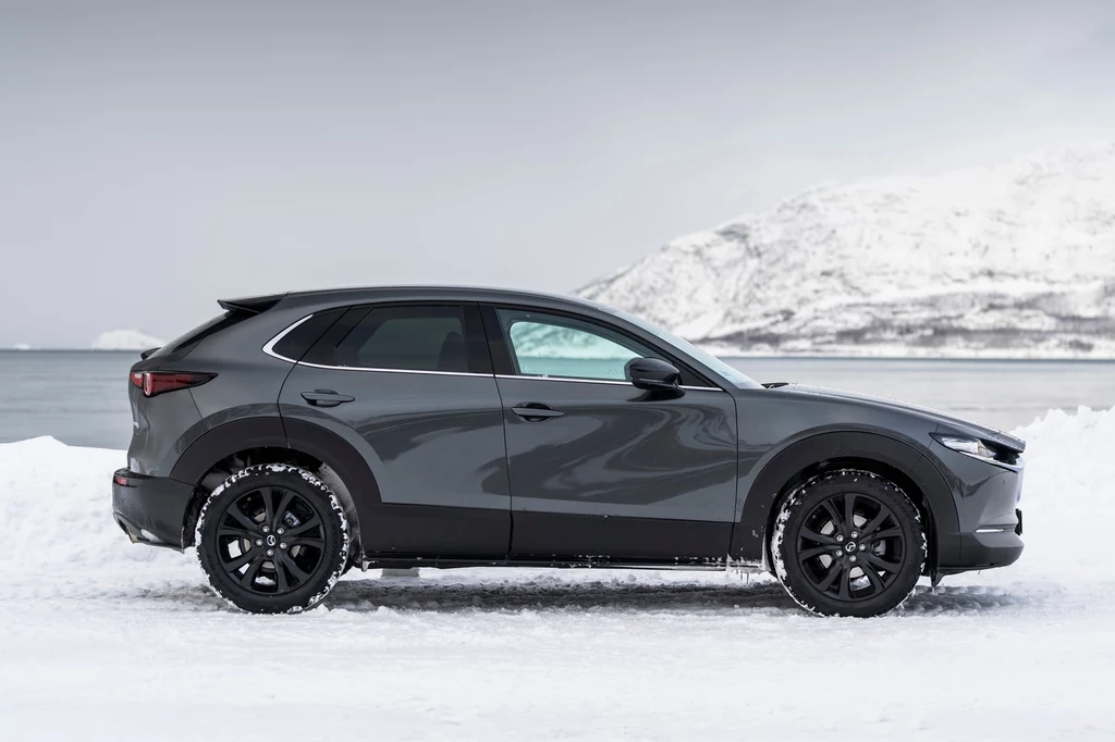 Mazda CX-30 to najchętniej kupowany model tej marki w Polsce Szare auto osobowe typu SUV stoi na pokrytej śniegiem drodze, w tle widoczne są zimowe wzgórza oraz zamarznięte jezioro lub morze.