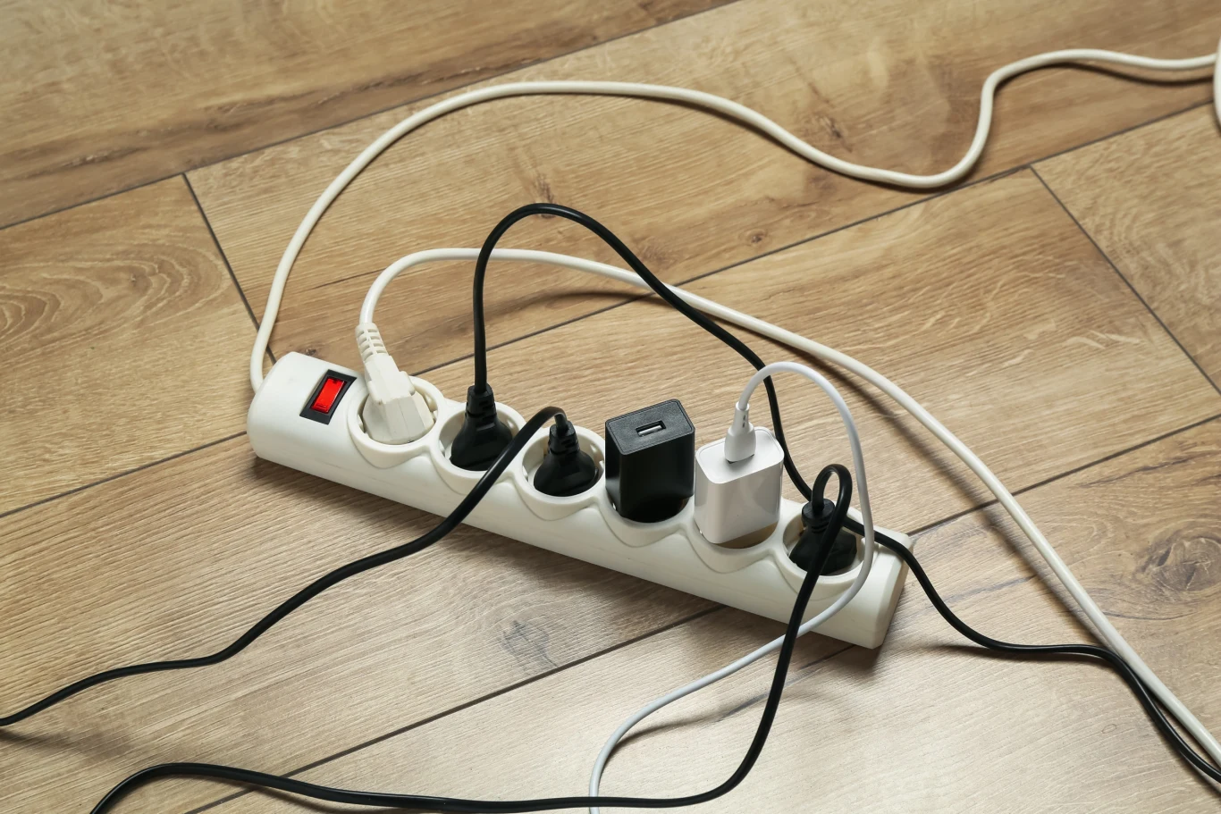 Pamiętaj o jednej rzeczy, a urządzenie elektryczne będą służyć dłużej Listwa zasilająca ustawiona na drewnianej podłodze jest podłączona do kilku różnych urządzeń za pomocą licznych kabli, wokół kabla widoczne są splątane przewody.