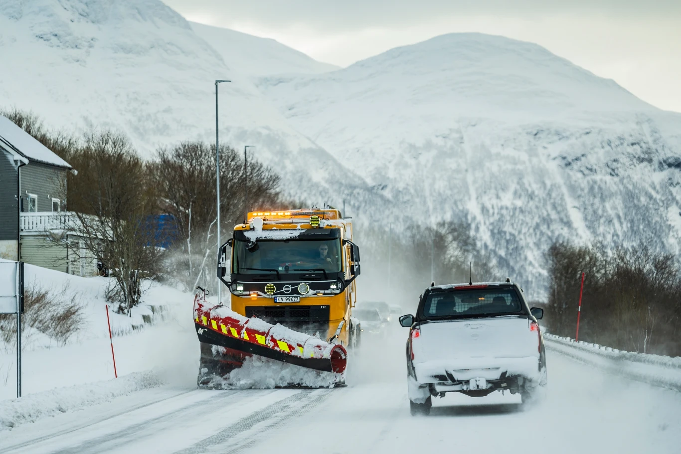 Ciężarówka odśnieżająca drogę podczas intensywnych opadów śniegu, obok przejeżdża samochód osobowy, w tle wysokie, ośnieżone góry i nieliczne drzewa przy drodze.