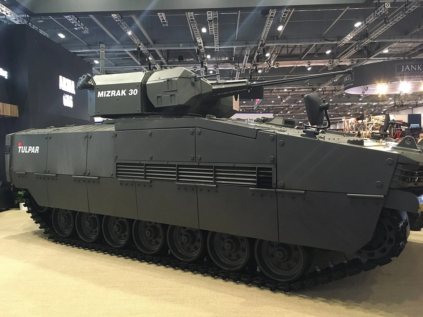 Ottokar Tulpra na targach zbrojeniowych DSEI 2019 w Londynie Opancerzony wóz bojowy na wystawie, wyposażony w wieżę MIZRAK 30 i gąsienice, oznaczony logo Tulpar, otoczenie targów lub hali wystawienniczej.