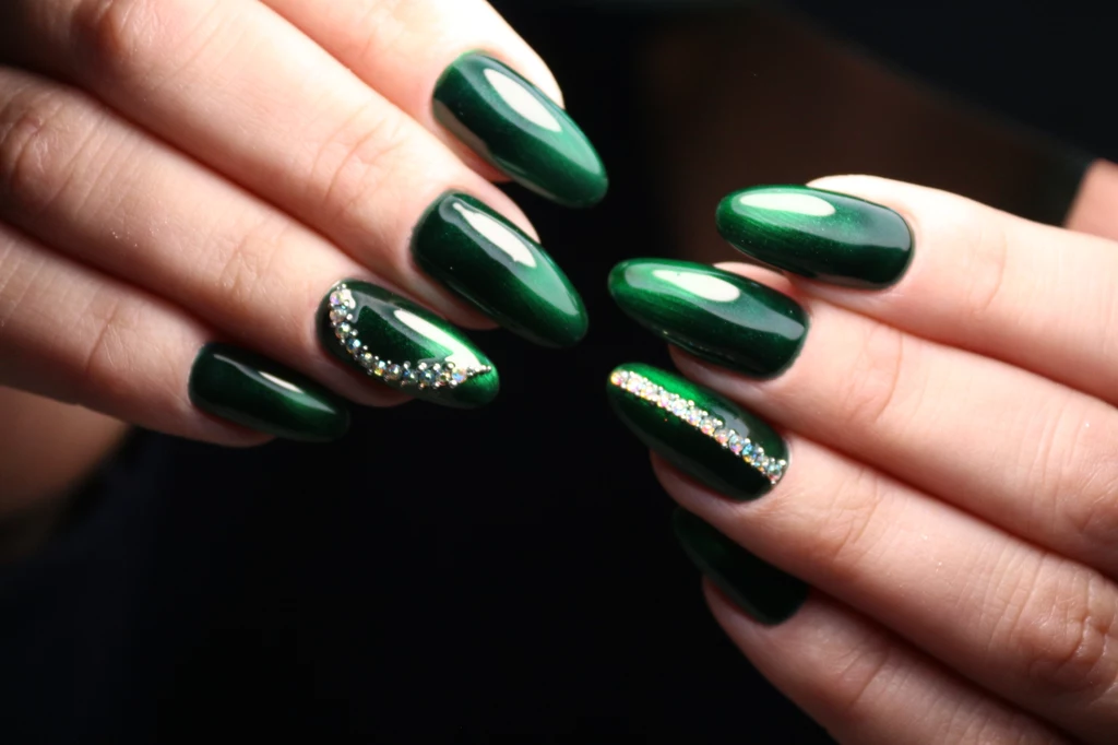 Szmaragdowy manicure świetnie pasuje zarówno na co dzień, jak i na wieczorne wyjścia Paznokcie pomalowane na ciemnozielony kolor z połyskującym wykończeniem, na dwóch paznokciach dekoracyjne aplikacje z kryształków i brokatu, dłonie w delikatnym, naturalnym układzie na ciemnym tle.