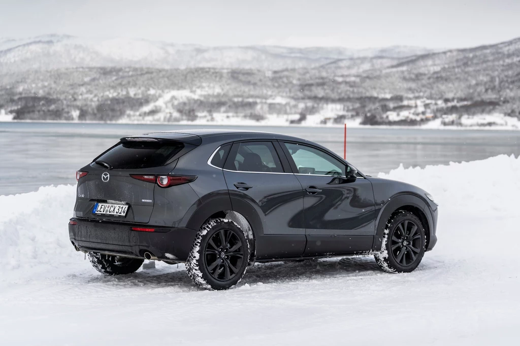 Aktualnie w polskiej ofercie modelu CX-30 znajduje się jedna wersja silnikowa, 2.0 e-Skyactiv-X Ciemnoszary samochód Mazda CX-30 stoi zaparkowany na zaśnieżonej drodze nad jeziorem, na tle ośnieżonych gór i spokojnej, zimowej scenerii.