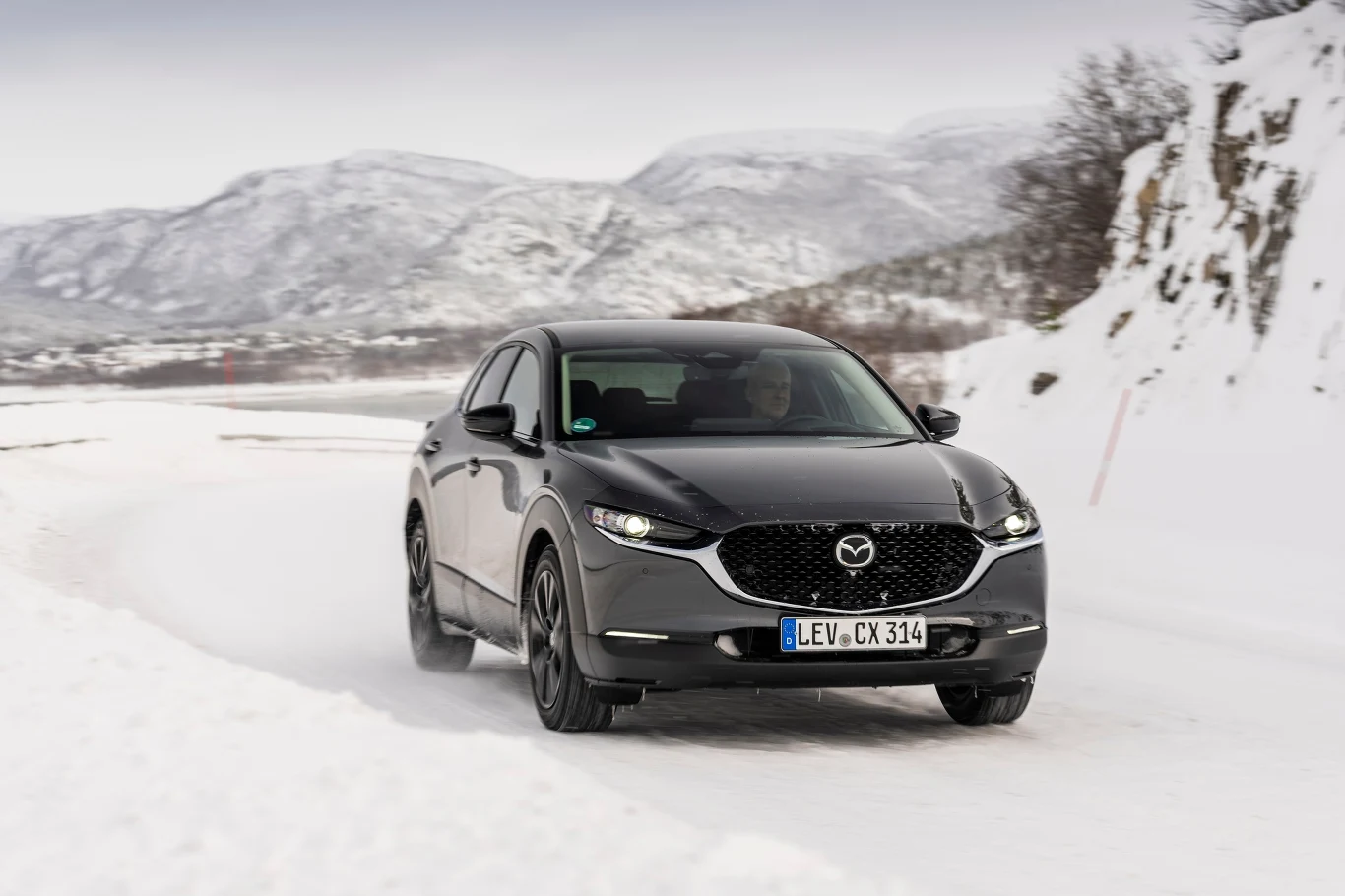 Czarny samochód marki Mazda jedzie po ośnieżonej, krętej drodze w górskim otoczeniu, a w tle widoczne są zasypane śniegiem pagórki i drzewa.