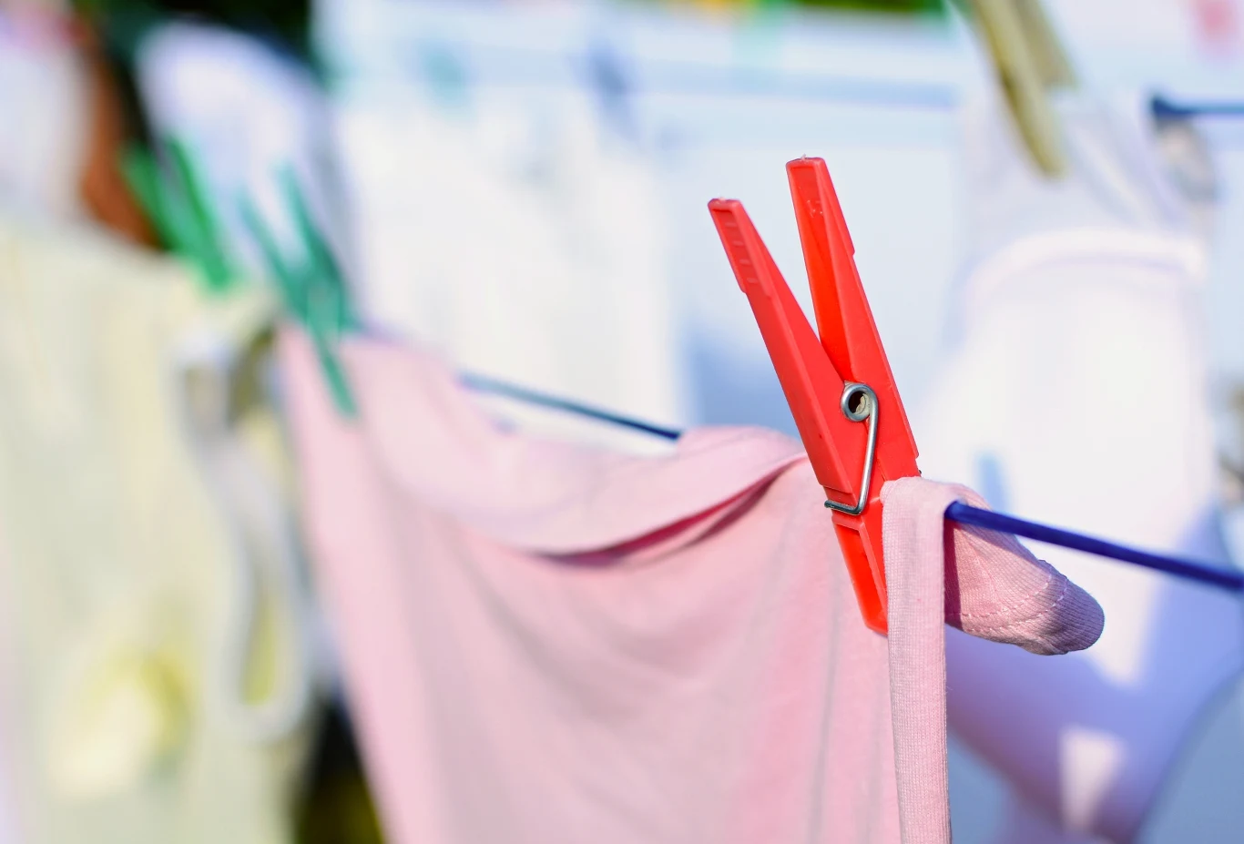 Jak dbać o bieliznę? Czerwona klamerka przypięta do różowego materiału wiszącego na sznurku na tle rozmytej bieli innych ubrań suszących się w słońcu.
