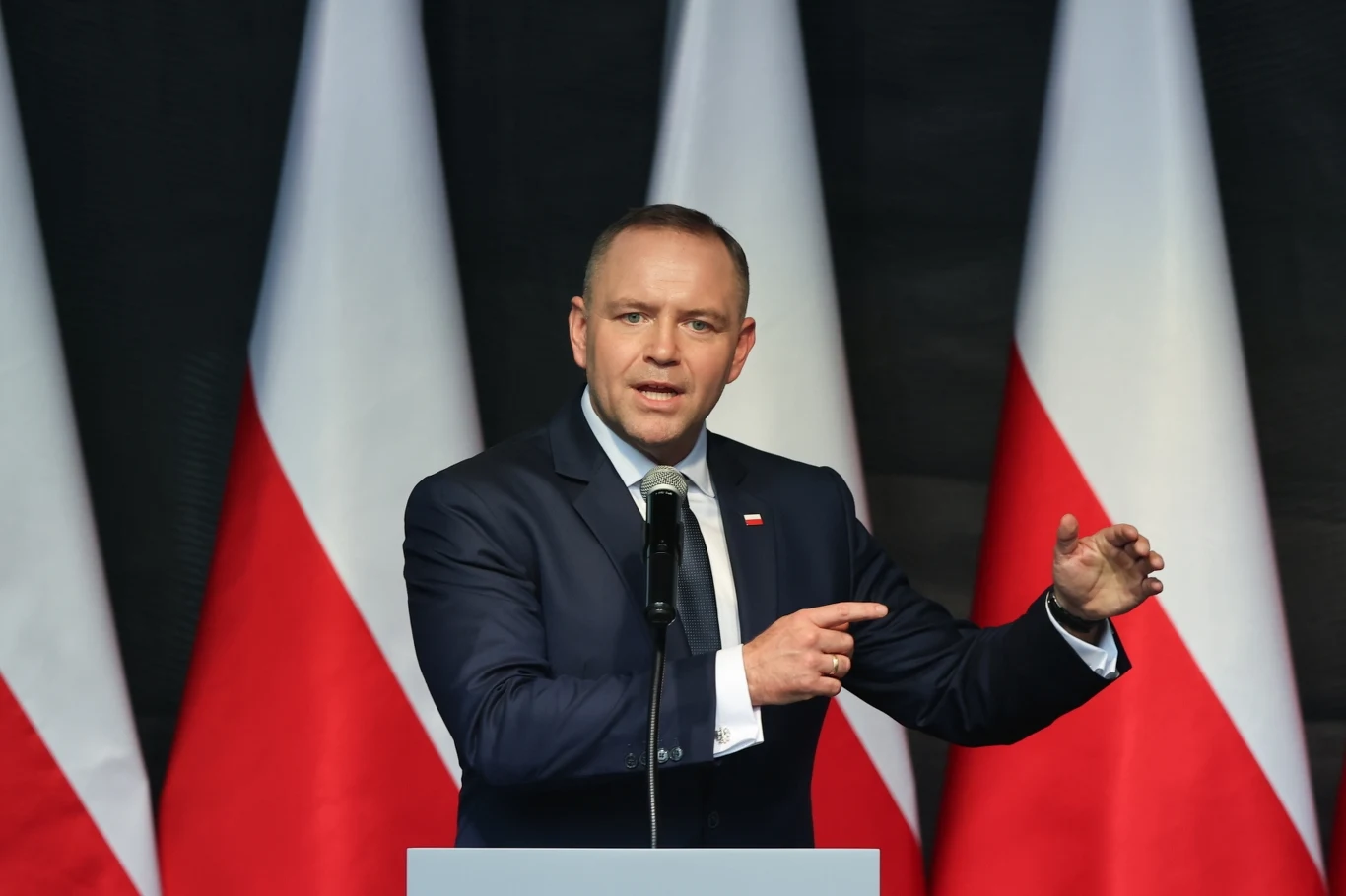 Prezydent Karol Nawrocki Mężczyzna w garniturze przemawia przy mównicy z mikrofonem, w tle biało-czerwone flagi narodowe.