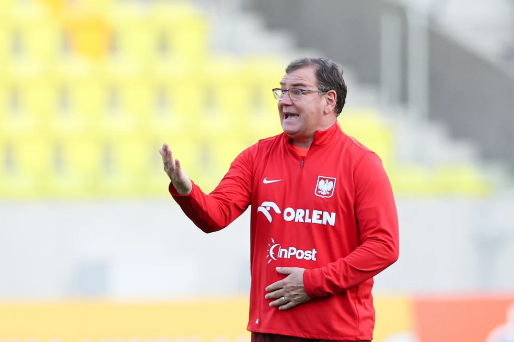 Jan Urban Trener w czerwonej bluzie z logo Polskiego Związku Piłki Nożnej i sponsora gestykuluje podczas meczu lub treningu na tle żółtych trybun.