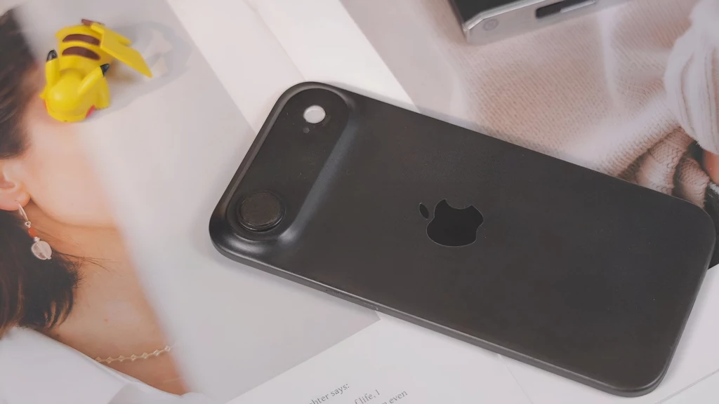 Makieta smartfona iPhone 17 Air. Czarny case na telefon z logo Apple leży na powierzchni pokrytej rozrzuconymi wydrukami zdjęć i dokumentów, obok znajduje się żółta figurka oraz fragment kobiecej twarzy widoczny na jednym z wydruków.
