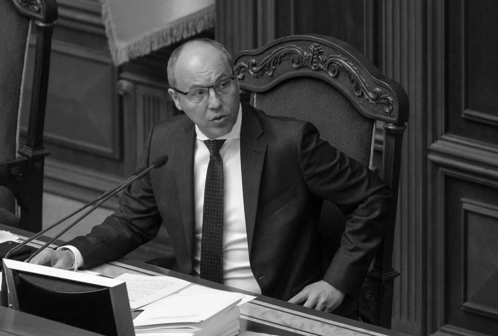 Były przewodniczący Rady Najwyższej Ukrainy Andrij Parubij Mężczyzna w garniturze siedzi przy biurku w eleganckim, drewnianym gabinecie, przed nim leżą stosy dokumentów, a on zwraca się w stronę mikrofonu jakby przemawiał.