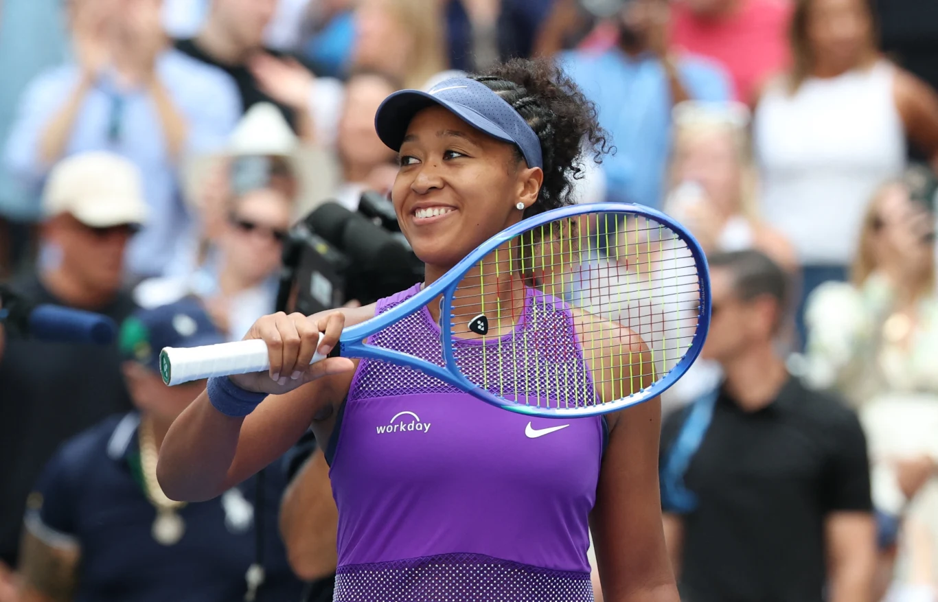 Naomi Osaka Tenisistka w fioletowym stroju z rakietą tenisową w ręku, uśmiechnięta, podczas meczu na korcie, w tle publiczność skupiona na wydarzeniu.