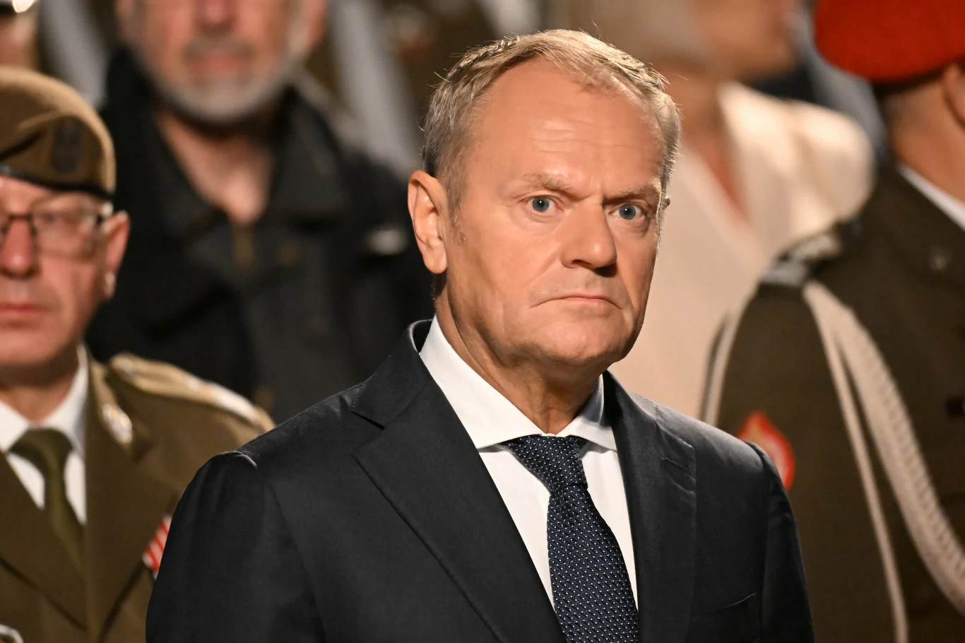 Donald Tusk Mężczyzna w garniturze i krawacie z poważnym wyrazem twarzy, otoczony osobami w wojskowych mundurach, w tłumie podczas oficjalnej uroczystości.