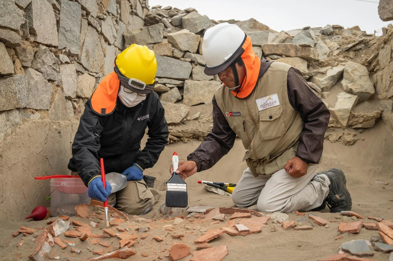Dwóch archeologów w ochraniaczach na głowie i twarzy pracuje przy wykopaliskach, starannie oczyszczając szczątki ceramiczne z piasku na tle kamiennych murów.