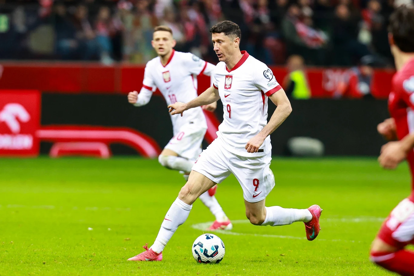 Robert Lewandowski i Sebastian Szymański w barwach reprezentacji Polski Polski piłkarz w białym stroju kontroluje piłkę podczas meczu, wokół niego znajduje się kilku innych zawodników w ruchu, stadion wypełniony kibicami w tle.